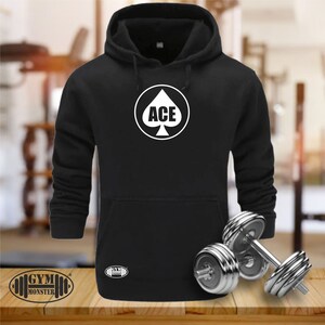 May include: Black hooded sweatshirt with a white graphic of a spade playing card with the word "ACE" inside. The sweatshirt is on a wooden surface with two silver dumbbells.