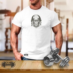 May include: White t-shirt with a graphic of a gorilla wearing a military helmet. The gorilla is looking forward with a serious expression. The t-shirt has the text "GYM MONSTER" printed on the bottom.