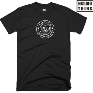 Op de afbeelding: Zwart T-shirt met een witte cirkelvormige afbeelding met de tekst "Vintage Est. 1973 Classic Edition A Legend Was Born"