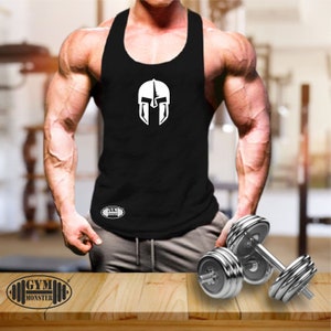 Könnte beinhalten: Schwarzes ärmelloses Tanktop mit einem weißen Spartanerhelm-Motiv und dem Text "GYM MONSTER" in einem weißen Oval. Das Tanktop wird von einer Person mit muskulösen Armen getragen.