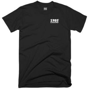 Op de afbeelding: Zwart T-shirt met witte tekst "1985 LEGEND" op de voorkant.