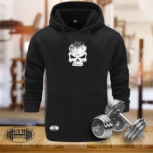 Op de afbeelding: Zwart hoodie met een witte schedelprint en een distressed print. De hoodie heeft een kangoeroezak en een capuchon met trekkoord. De tekst "GYM MONSTER" is gedrukt in de linkerbenedenhoek van de afbeelding.