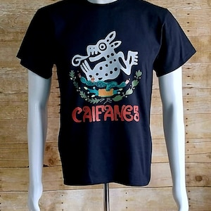 May include: Black t-shirt featuring a white and gray illustration of a frog-like creature with the word "CAIFANES" in red. The design includes a wreath and other graphic elements. The shirt is displayed on a mannequin.