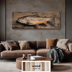 Op de afbeelding: Een houten wandkunstwerk met een gedetailleerde illustratie van een bruine forel. De vis is afgebeeld met realistische kleuren en textuur, gemonteerd op een rustieke houten plank achtergrond. Het kunstwerk wordt opgehangen door een dunne draad. De tekst "wood wall art" staat onderaan.