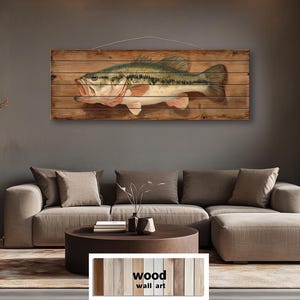 Op de afbeelding: Houten wandkunst met een gedetailleerde illustratie van een largemouth bass. De vis is afgebeeld met groene, witte en roze tinten, gemonteerd op een rustieke houten plank achtergrond. Het kunstwerk is ontworpen voor wanddecoratie.