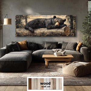 May include: Wood wall art featuring a black bear resting on a log. The artwork is mounted on a wooden plank background, creating a rustic aesthetic. The bear is depicted in a realistic style, with detailed fur and a peaceful expression. The artwork is displayed in a living room setting.