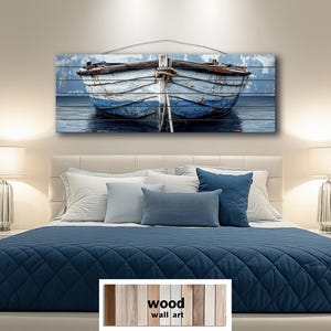 Op de afbeelding: Houten wandkunst met een verweerde witte en blauwe boot op een blauwe waterachtergrond. De boot is gecentreerd en vastgebonden met een touw. De kunst is gemonteerd op houten planken, wat een rustieke uitstraling creëert.