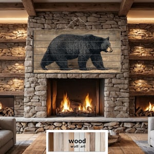 May include: A black bear walking on a wooden panel, mounted above a fireplace. The artwork is displayed in a rustic living room with stone walls and wooden beams. The words "wood wall art" are visible.
