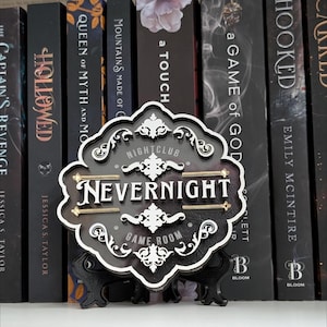 May include: A black and white decorative sign with the text "NEVERNIGHT NIGHTCLUB GAME ROOM" displayed in a stylized font. The sign is placed in front of a row of books with dark covers. The sign is supported by a black stand.