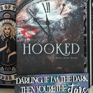 May include: The book cover for "HOOKED: A Never After Novel" displays a broken clock face design with red details and the title in silver. A decorative sign below reads "DARLING, IF I'M THE DARK THEN YOU'RE THE stars" in white lettering.