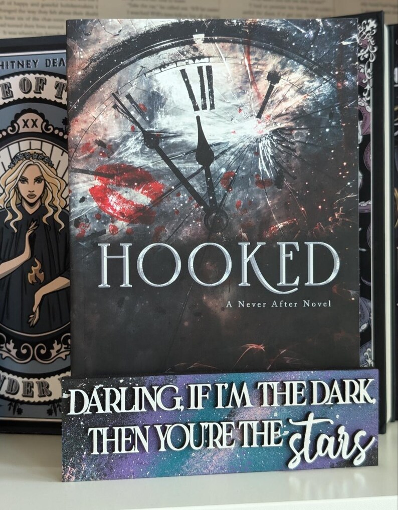May include: The book cover for "HOOKED: A Never After Novel" displays a broken clock face design with red details and the title in silver. A decorative sign below reads "DARLING, IF I'M THE DARK THEN YOU'RE THE stars" in white lettering.