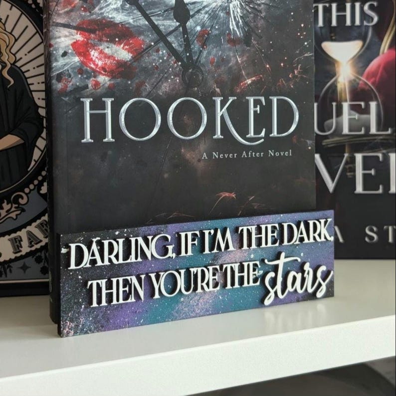 May include: A wooden sign with the quote "DARLING IF I'M THE DARK THEN YOU'RE THE stars" in white lettering. The sign has a galaxy-themed background with purple, blue, and white colours. A book with the title "HOOKED" is behind the sign.