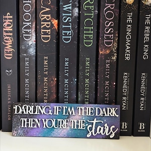 May include: A selection of hardback books with titles including "Hooked," "Scarred," and "Twisted." A decorative sign with the text "Darling, if I'm the dark, then you're the stars" is in front of the books. The books have a dark background with colourful accents.