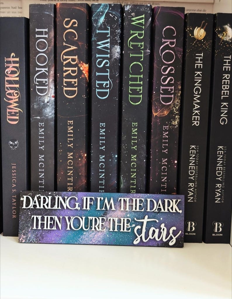 May include: A selection of hardback books with titles including "Hooked," "Scarred," and "Twisted." A decorative sign with the text "Darling, if I'm the dark, then you're the stars" is in front of the books. The books have a dark background with colourful accents.