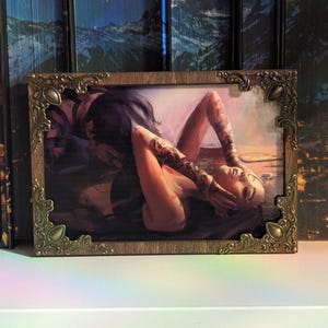 May include: A rectangular art print featuring a digital illustration of two figures in an intimate embrace, enclosed in a dark wood-toned frame with ornate, antique-style bronze metal corner accents that feature teardrop-shaped details.