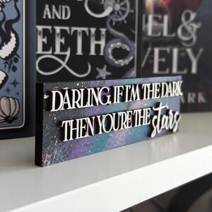 May include: A wooden sign with the quote "DARLING, IF I'M THE DARK THEN YOU'RE THE STARS" in white, raised lettering. The background features a galaxy design in purple, blue, and white. Other books are visible in the background.