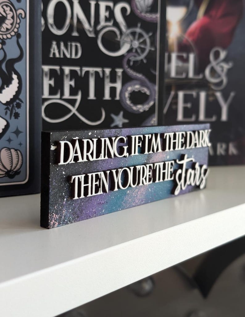May include: A wooden sign with the quote "DARLING, IF I'M THE DARK THEN YOU'RE THE STARS" in white, raised lettering. The background features a galaxy design in purple, blue, and white. Other books are visible in the background.