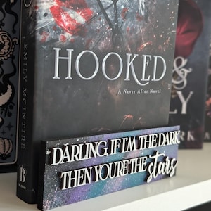 May include: A book titled "HOOKED" with a decorative wooden sign. The sign displays the quote "DARLING, IF I'M THE DARK THEN YOU'RE THE STARS" in white lettering against a purple and blue space-like background.