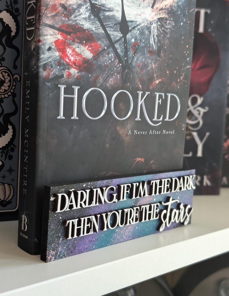 May include: A book titled "HOOKED" with a decorative wooden sign. The sign displays the quote "DARLING, IF I'M THE DARK THEN YOU'RE THE STARS" in white lettering against a purple and blue space-like background.