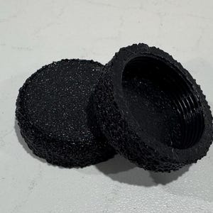 May include: Two black, textured, round objects. One is a solid disc, and the other is a hollow, cup-like shape with interior threading. The objects have a rough, speckled surface.