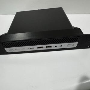 May include: Black computer case with a silver front panel. The front panel has the text "ELITEDESK" and various ports. The case is mounted in a black bracket with mounting holes on each side. A black device sits on top of the case.