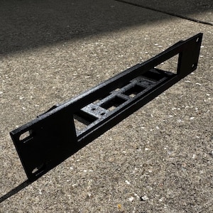 May include: A black metal rack mount component with rectangular and square cutouts. The component has a textured surface and is designed for mounting equipment. The item is angled, casting a shadow on the surface.