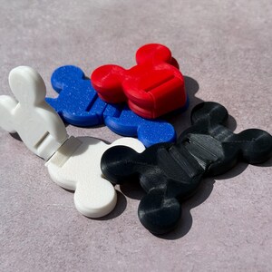 May include: A collection of colorful, Mickey Mouse-shaped cable organizers. The organizers are in white, blue, red, and black. Each has a slot for holding cables. The items are made of plastic.