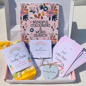 May include: A gift box with a colouring book, socks, a bracelet, and cards. The colouring book is titled "Mindful Colouring & Word Search". The socks are yellow with pineapple designs and the text "IVF Lucky Socks". The bracelet and cards are labeled "IVF Little Steps".
