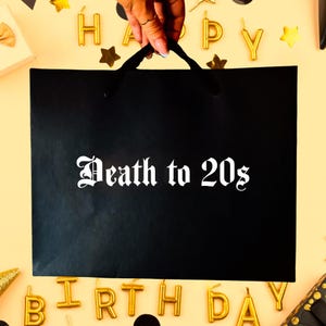 May include: A black gift bag with the text "Death to 20s" in white gothic font. The bag is held by a hand. Gold "Happy Birthday" and star-shaped decorations surround the bag.