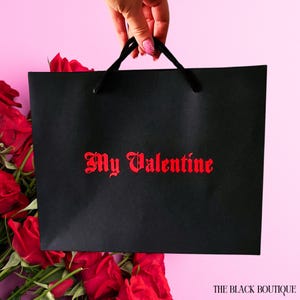 May include: A black gift bag with the words "My Valentine" in red script. The bag is held by a hand with pink nail polish. Red roses are in the background against a pink backdrop. The Black Boutique logo is in the bottom right corner.