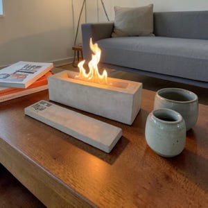 May include: A rectangular concrete fireplace with a flame burning inside. A concrete lid is in front of the fireplace. Two small, light green ceramic cups are to the right. The items are on a dark wood table.
