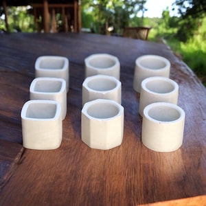 Puede incluir: Nueve pequeñas macetas de hormigón gris claro con diversas formas geométricas. Algunas son rectangulares, otras octogonales, todas con la parte superior abierta. Están dispuestas sobre una superficie de madera oscura, para pequeñas plantas o suculentas.