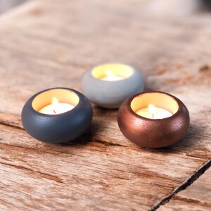 Peut inclure: Trois petits bougeoirs ronds de couleurs bleu, gris et bronze, chacun avec une bougie chauffe-plat allumée à l'intérieur. Les bougeoirs sont disposés sur une surface en bois rustique, créant une ambiance chaleureuse.