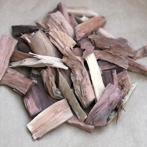 May include: A collection of wood chips in shades of brown and beige. The chips have a rough, textured appearance, with visible grain and splintered edges. The wood pieces are scattered on a light brown surface.