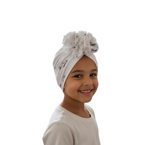 May include: A white baby turban with a floral accent. The turban has a subtle pattern of black shapes. The floral accent is made of ruffled fabric. The turban is worn by a young child.