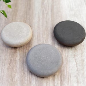 May include: Three smooth, round, stone coasters in varying shades of gray and beige. The coasters are arranged on a light wood-grain surface, with a few green leaves in the upper left corner.