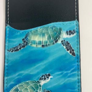 May include: Black leather card holder with a blue and green sea turtle design. The card holder has a stitched edge and a flap that opens to reveal a pocket for cards.