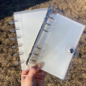 May include: Two clear, white, ring binder notebooks with silver metal rings and a snap closure. The notebooks are held open, revealing the interior pages. The background is blurred, with a hint of green grass.