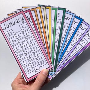 May include: A hand holds a fan of colorful savings tracker cards, each representing a month. The cards have a white background with a month's name and boxes with amounts like $2, $4, and $50. The edges are outlined in various colors.