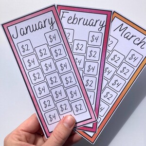 May include: Three colorful savings tracker cards for January, February, and March. Each card has a different colored border and features a grid of boxes with dollar amounts. The cards are held in a hand.