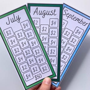 May include: Three colorful savings tracker cards for July, August, and September. Each card has a different colored border and features a grid of boxes with dollar amounts. The cards are held by a hand.