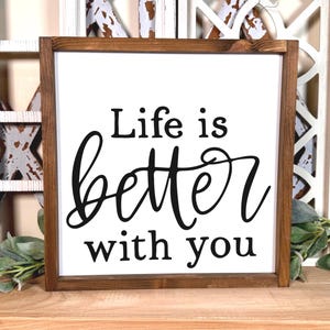 May include: A square wooden sign with a dark brown frame and a white background. The text "Life is better with you" is printed in black, featuring a combination of a simple sans-serif font and a decorative cursive script for the word better.