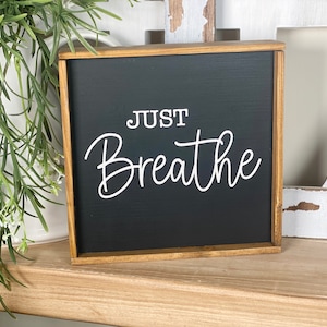 May include: A square wooden sign with a black background and a natural wood frame. The text "JUST Breathe" is printed in white in a combination of block and script lettering. The sign is positioned upright with a clean, simple design.