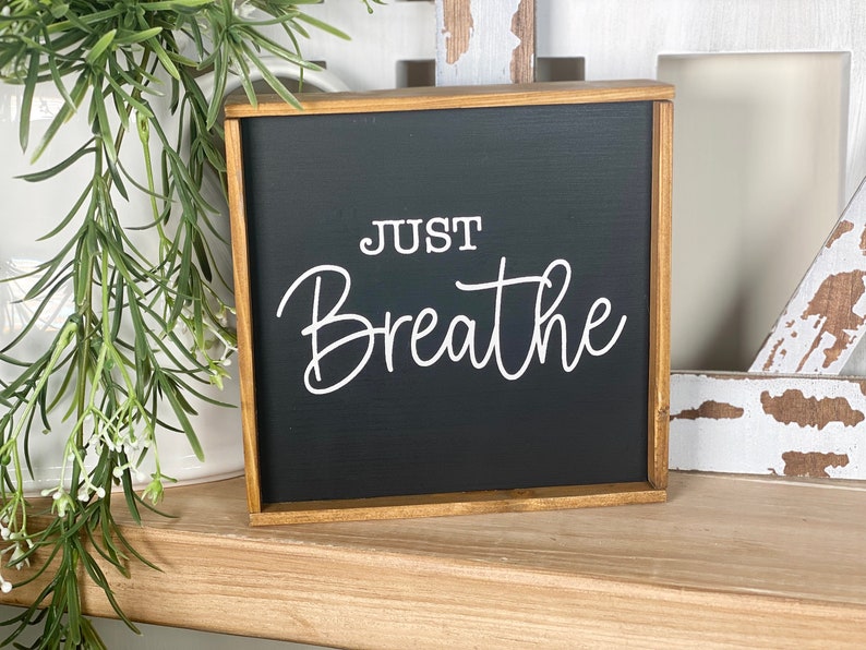 May include: A square wooden sign with a black background and a natural wood frame. The text "JUST Breathe" is printed in white in a combination of block and script lettering. The sign is positioned upright with a clean, simple design.