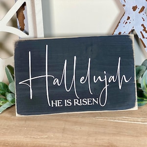May include: A rectangular, dark gray wooden sign with the word "Hallelujah" in white cursive script. Below, the text "He is Risen" is printed in a smaller, sans-serif font. The sign has a distressed, rustic appearance.
