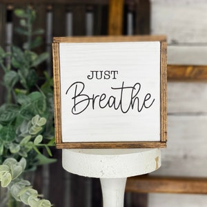 May include: A square wooden sign with a white face and dark brown stained edges. The text "JUST Breathe" is printed in black, with "JUST" in a simple sans-serif font and "Breathe" in a flowing script font. The sign has a visible wood grain.