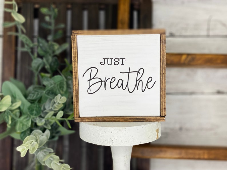 May include: A square wooden sign with a white face and dark brown stained edges. The text "JUST Breathe" is printed in black, with "JUST" in a simple sans-serif font and "Breathe" in a flowing script font. The sign has a visible wood grain.
