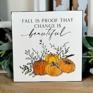 May include: A square, white wooden sign with the text "FALL IS PROOF THAT CHANGE IS beautiful" in black script. The sign features a watercolor illustration of four pumpkins in shades of orange and yellow.