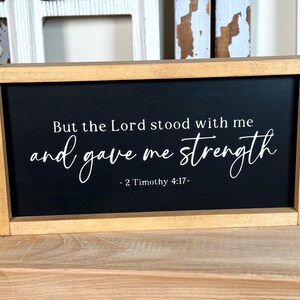May include: A rectangular wooden sign with a black face and white text. The text reads "But the Lord stood with me and gave me strength - 2 Timothy 4:17-". The frame is light brown.