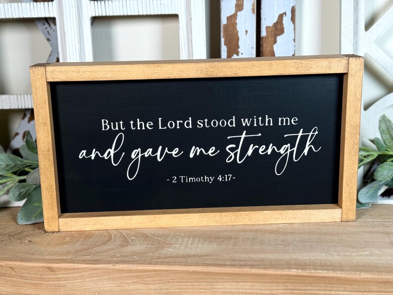 May include: A rectangular wooden sign with a black face and white text. The text reads "But the Lord stood with me and gave me strength - 2 Timothy 4:17-". The frame is light brown.